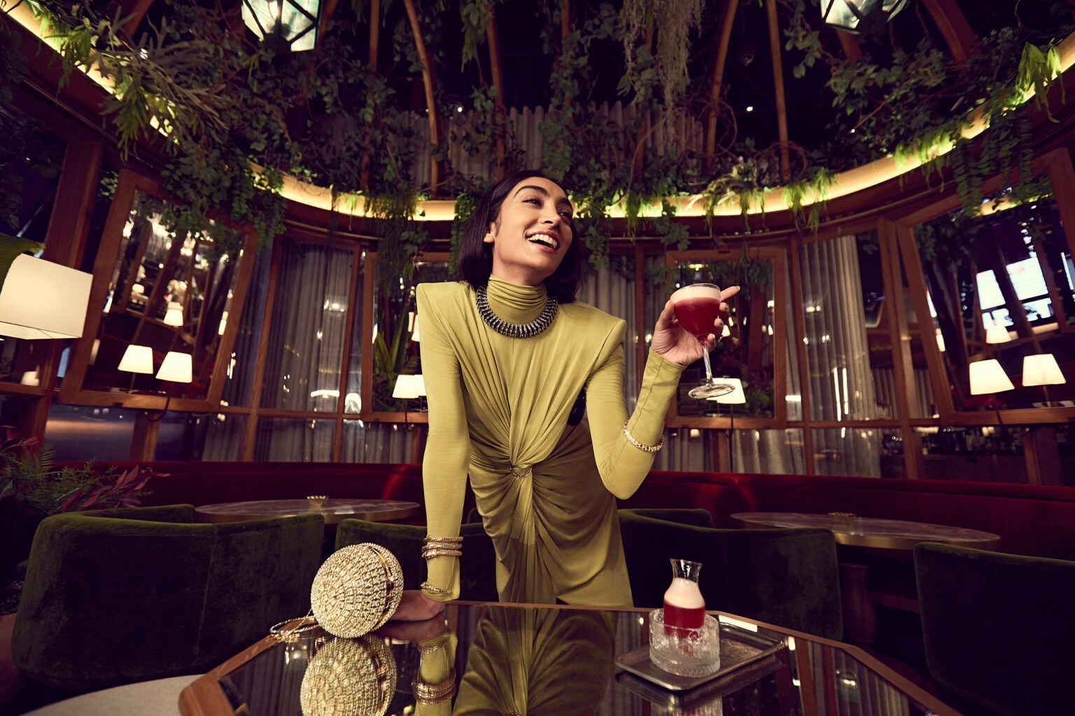 Woman raising a cocktail in a candlelit lounge with a leafy glass ceiling