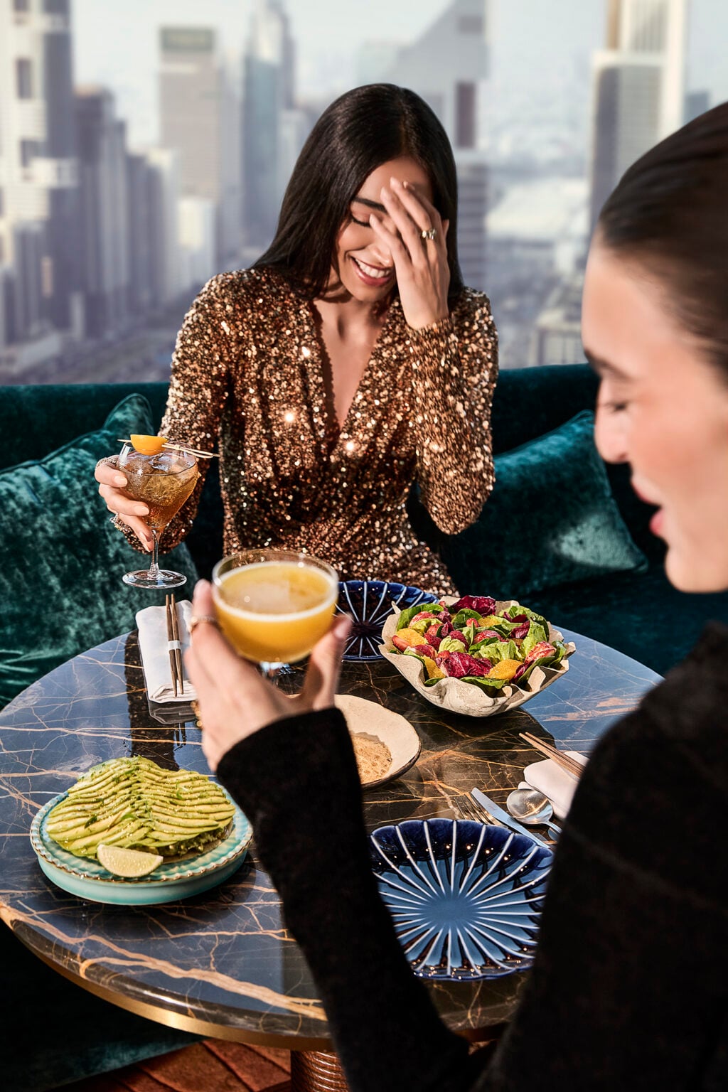 Woman laughing with a cocktail at a marble table above the Dubai skyline