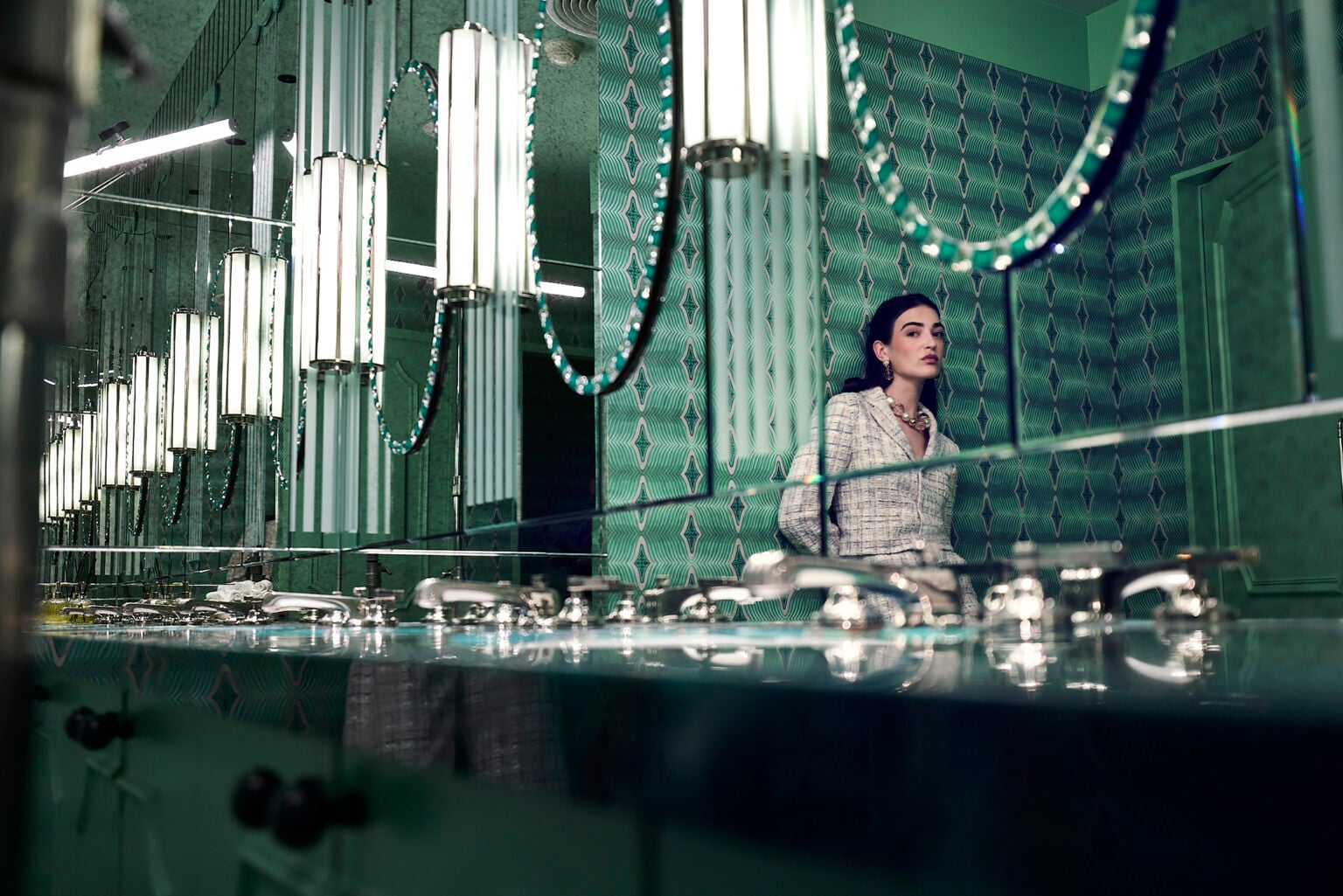 Woman reflected in the mirrors of a green-tiled washroom with glass pendant lights
